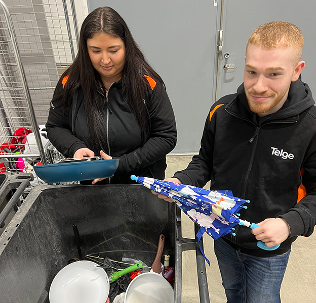 Två personer står vid en avfallsbinge och håller i en stekpanna och ett paraply